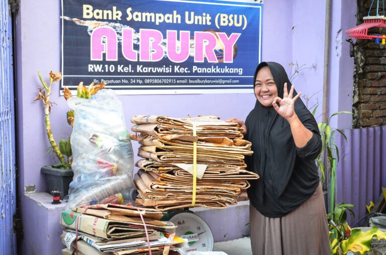 A woman smiles with her hand atop a stack of flattened cardboard in front of a purple wall.