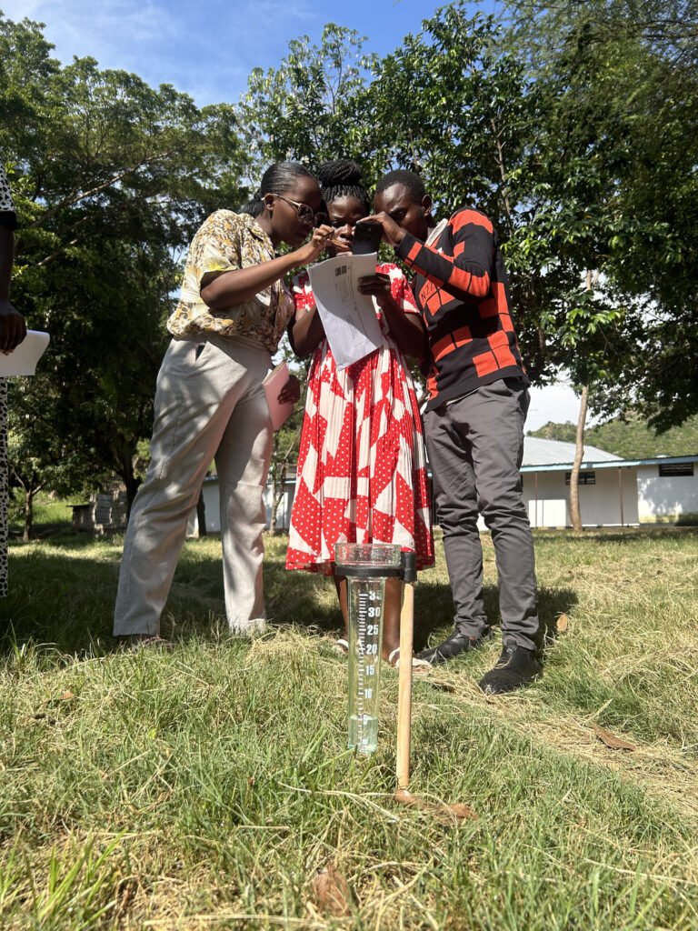 Three people consult a piece of paper behind a rainfall measurement tube and stick in the grass.