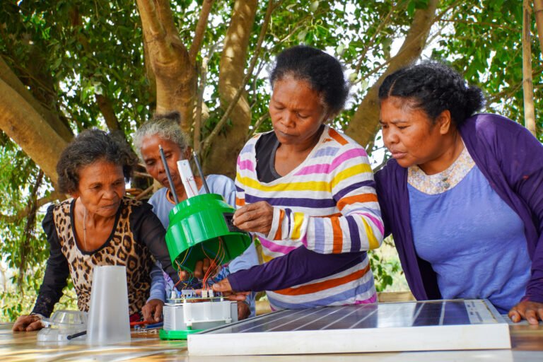 Four women connect electrical wires to a solar panel in an outdoor workspace.