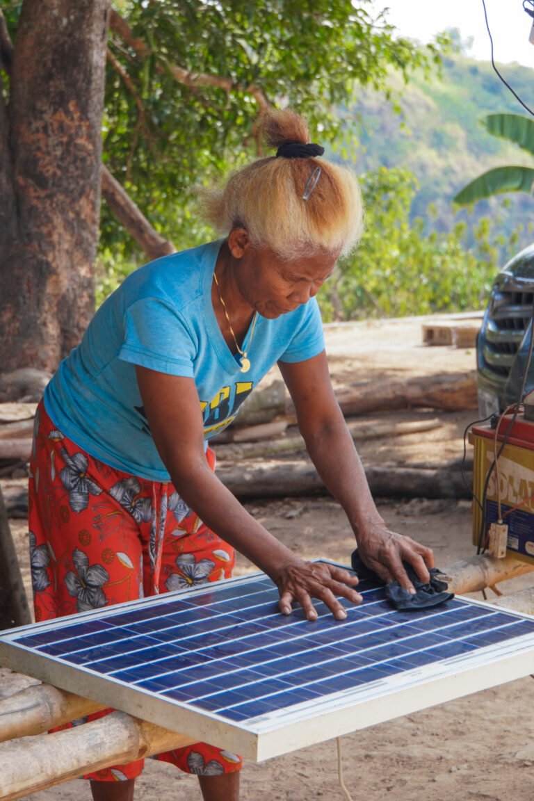 A woman bends over to clean a solar panel laid across tree branches.
