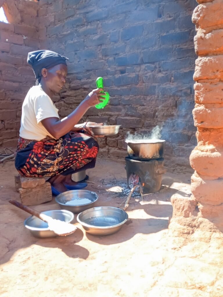A woman sits on a dirt floor in a brick structure surrounded by cooking equipment and using a cookstove.