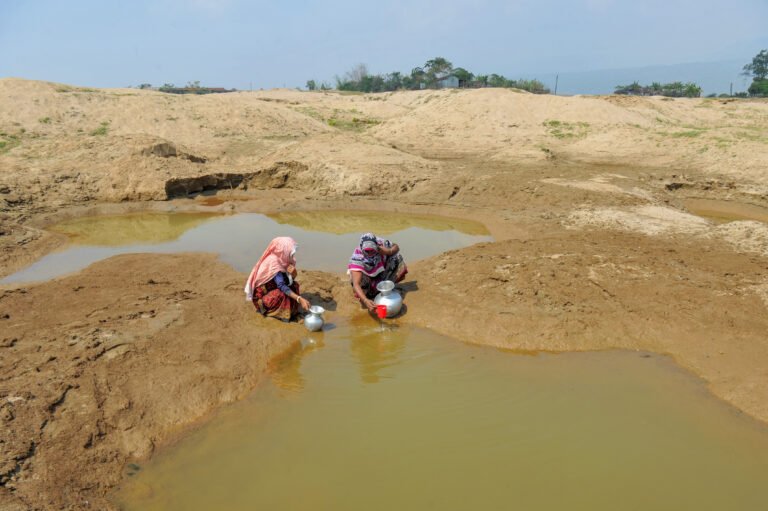 Two people collect water from a low reservoir in a dry landscape.