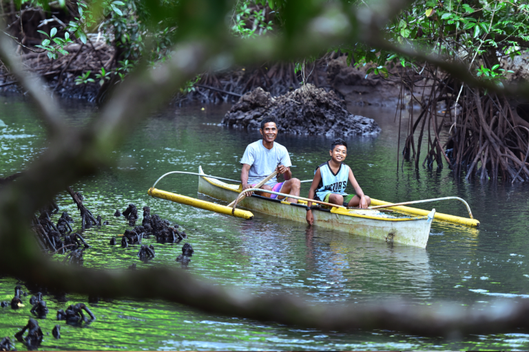A father and young adolescent son smile from a canoe as they paddle on a small river through a mangrove forest.