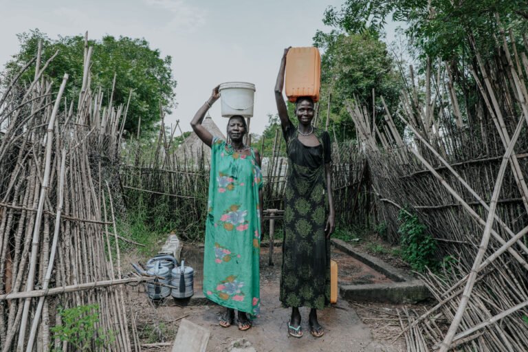 Two women standing in the Gambella region of Ethiopia, each carrying a water container on their head. They are in a pathway surrounded by bamboo fences with trees in the background. This scene illustrates the impact of the SunWASH project, which ensures access to clean drinking water for local communities.
