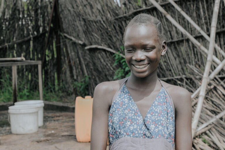 A young girl in the Gambella region of Ethiopia smiling, standing in front of a bamboo fence. She is happy because she no longer has to travel far to fetch clean water, allowing her to attend school without interruption and improving her safety against gender-based violence.