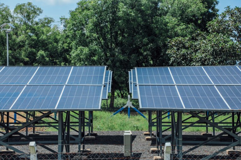 Solar panels installed on metal frames within a fenced area in a refugee camp in the Gambella region of Ethiopia. This solar-powered water system is part of the SunWASH project, providing clean drinking water to the community. Trees and a building are visible in the background under a partly cloudy sky.