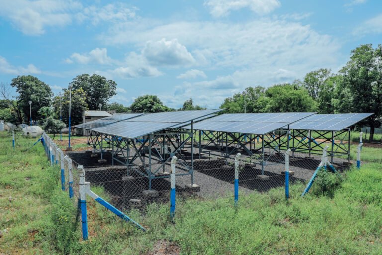 Solar panels installed on metal frames within a fenced area in a refugee camp in the Gambella region of Ethiopia. This solar-powered water system is part of the SunWASH project, providing clean drinking water to the community. Trees and a building are visible in the background under a partly cloudy sky.
