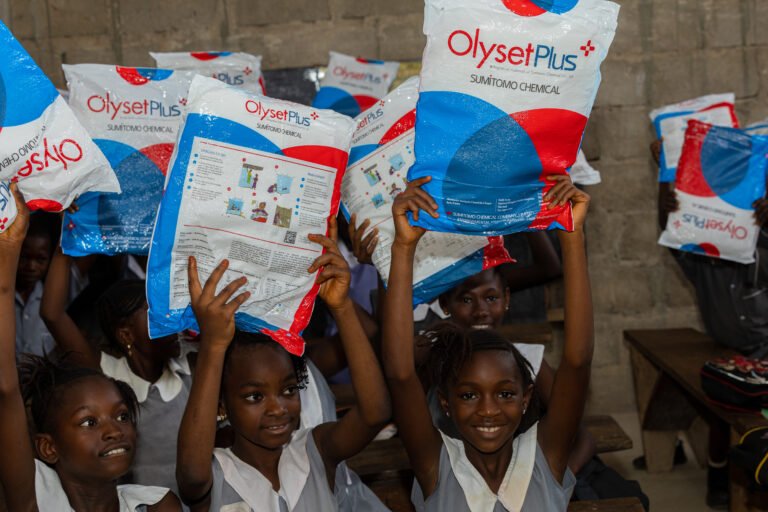 A group of school children hold packages of insecticide-treated nets above their heads while smiling.