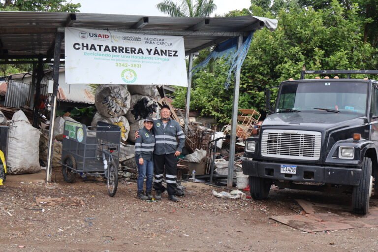 Two recycling collectors stand outside their truck under a canopy structure.