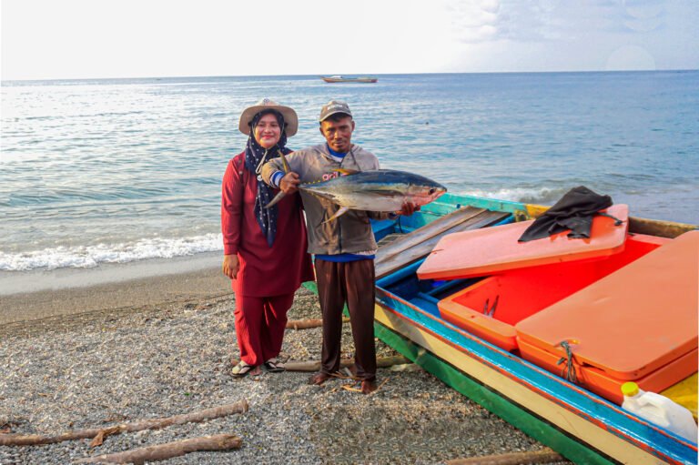 A woman smiles next to a man holding a large fish near a boat on the shore of the ocean.