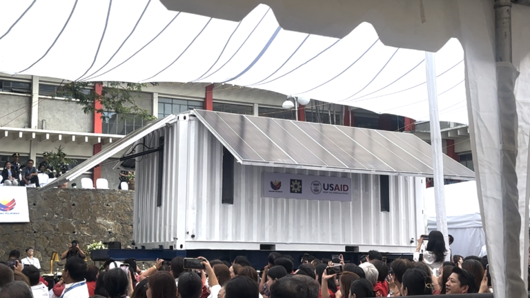A crowd gathers around a mobile building outfitted with solar panels.