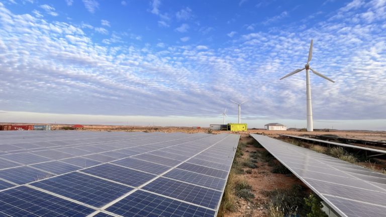 Rows of solar panels in a field with three wind turbines, a green electric generator, a cargo container and a building in the background against a blue sky with clouds