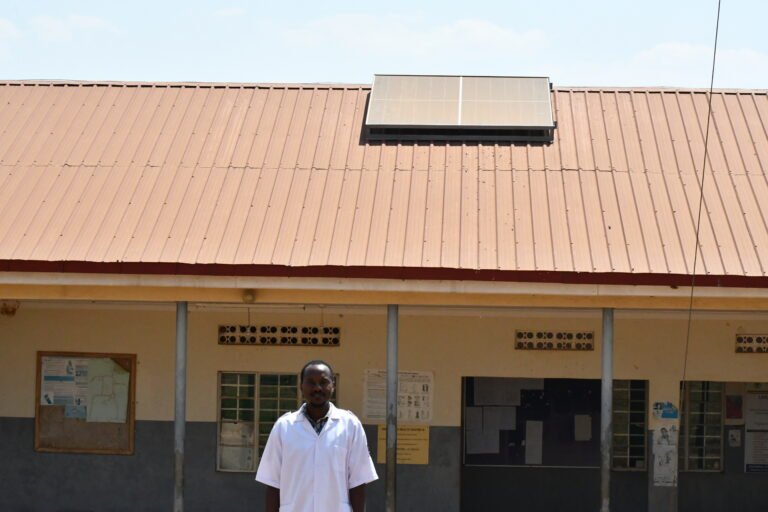 Martin standing infront of Lake Mburo Health Center III with solar panel on roof in the background