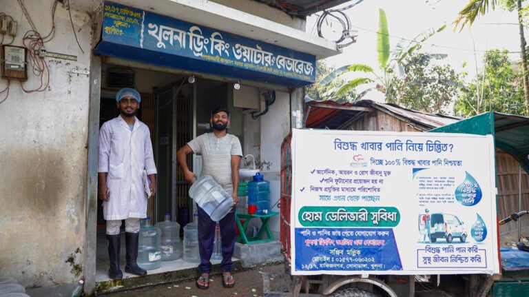 Two water delivery workers smile outside a home, with one carrying a 5-liter jug of drinking water.