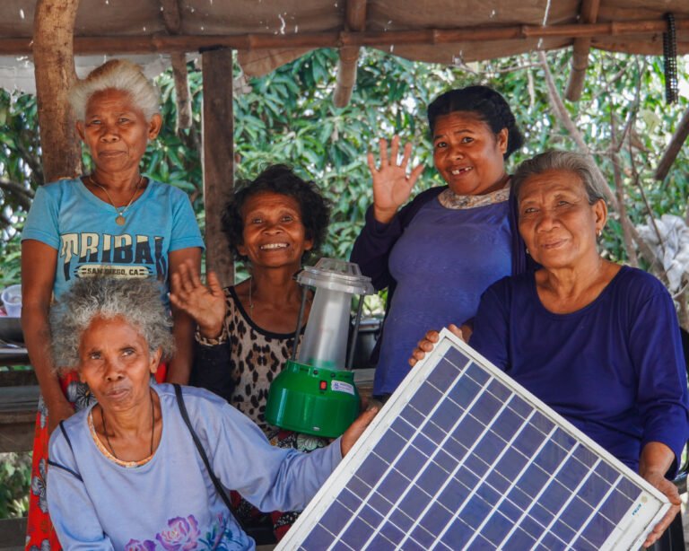 Five women smile and wave at the camera as they hold up a solar panel.