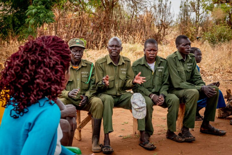 A group of men in scout uniforms sit side-by-side as one gestures to a woman sitting with her back turned to the camera, in a dry outdoor setting.