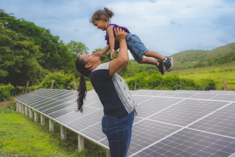 A woman lifts her child up next to solar panels with mountains in the background.