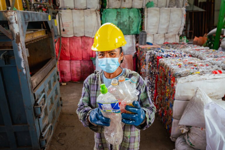 A woman wearing a mask and a hard hat holds up empty plastic bottles in a recycling facility.