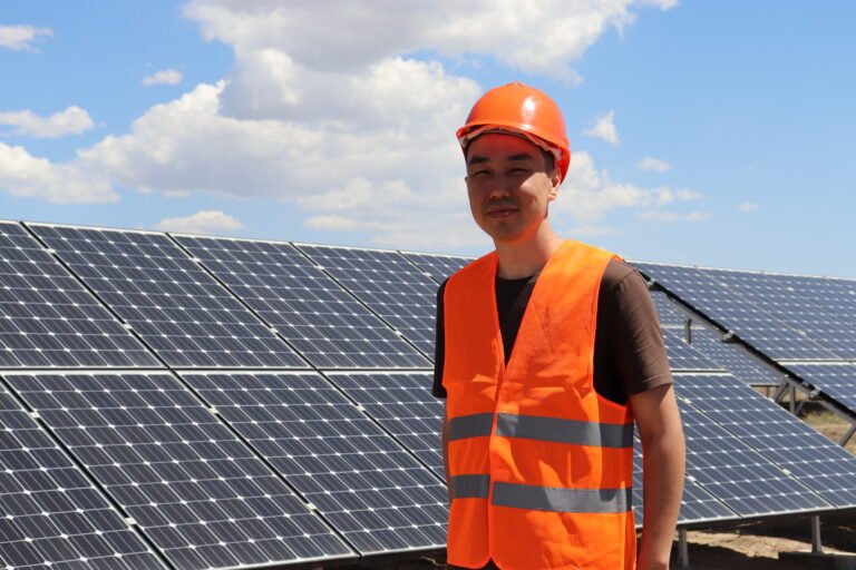 A man in an orange hard hat and safety vest smiles in front of solar panels outside.