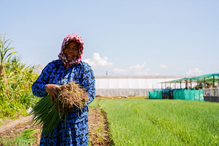 A woman smiles as she carries spring onions harvested from the farm behind her.