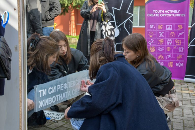 Two young women kneel down in front of a large mirror with a message written on it.