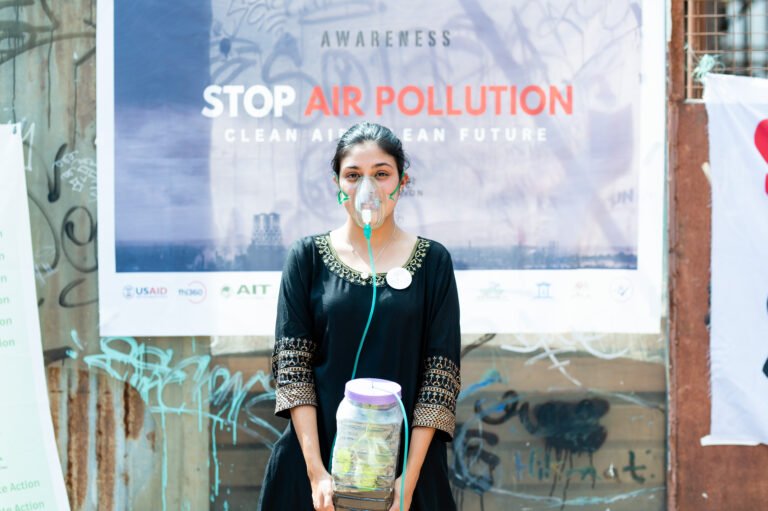 A young woman wears an oxygen mask drawing air from a plant enclosed in a container, facing the camera with a “Stop Air Pollution” sign behind her.