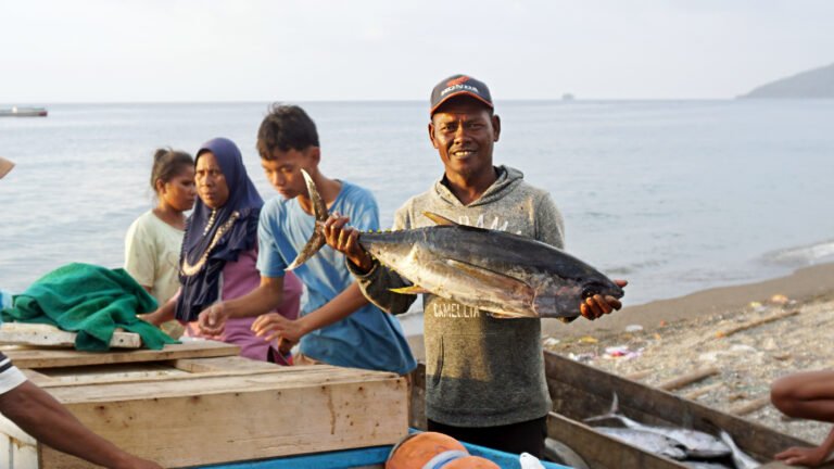 A man holds up his fish catch as three people work behind him, with the ocean in the background.
