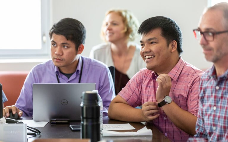 A group of people in a conference room, with the focus on two men in the center - one is on his laptop and one is laughing.