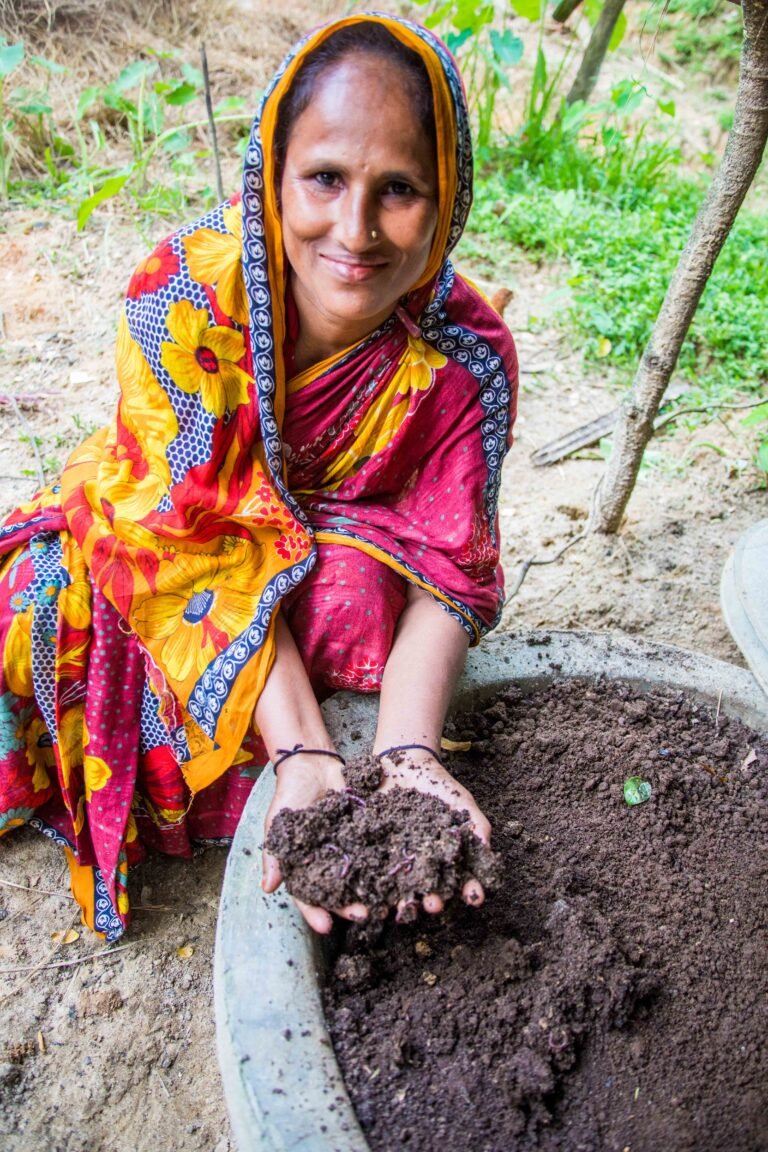 A woman in a colorful dress kneels and cups a handful of dark brown compost while smiling for the camera.