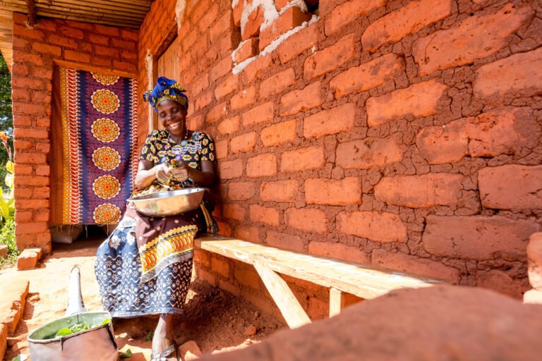 A woman in colorful traditional clothing sits in the shade of her house's patio, cleaning vegetables.