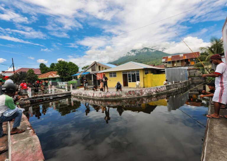 Colorful houses are surrounded by a small pond with fishermen fishing from the bank.