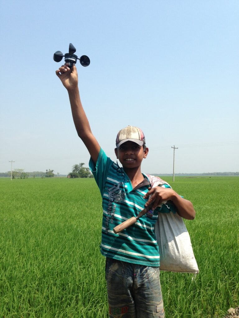 A boy wearing a cap holds up a cup anemometer in front of a green field and blue sky.