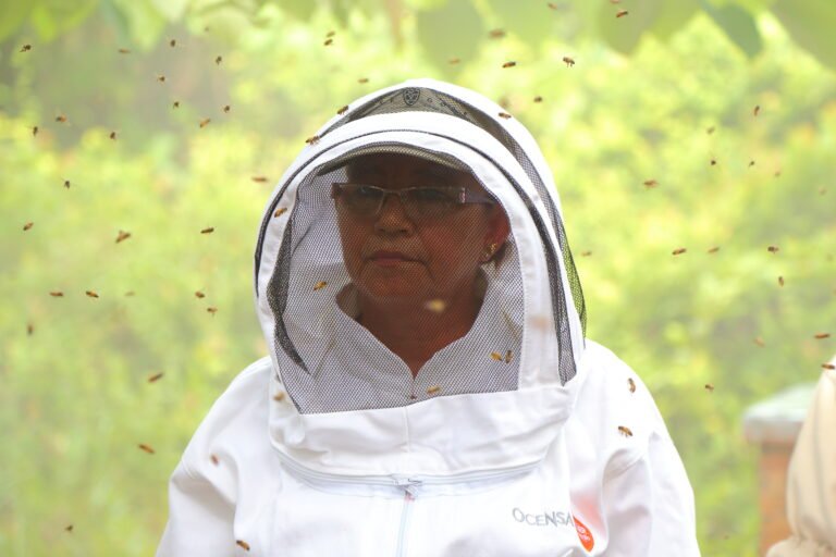 A woman wearing a beekeeper suit stands among many flying bees.
