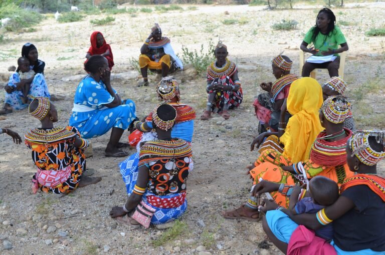 A group of women in colorful dresses sit in a loose circle and engage in discussion with each other.