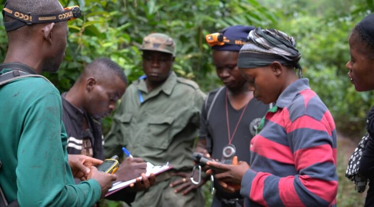 A group of people stand in a close circle filling out clipboards and looking at a GPS receiver.