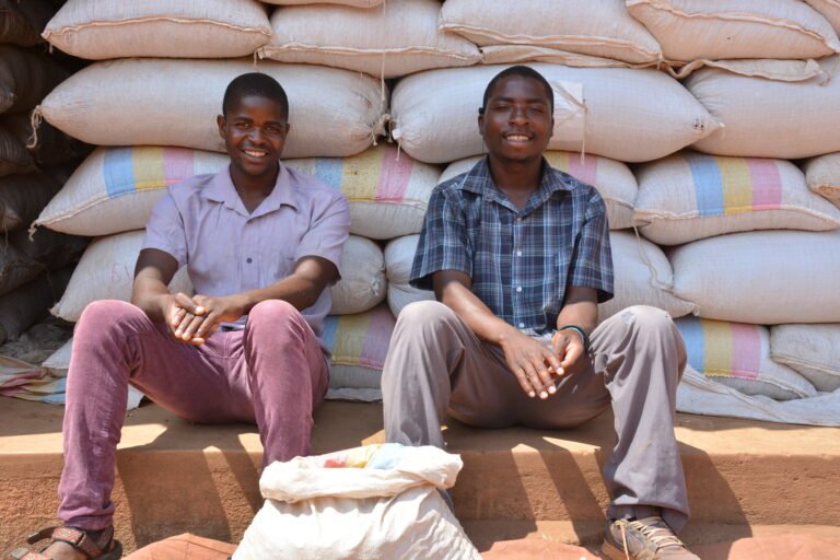 Two men sit in front of a stack of large grain bags, grinning at the camera.