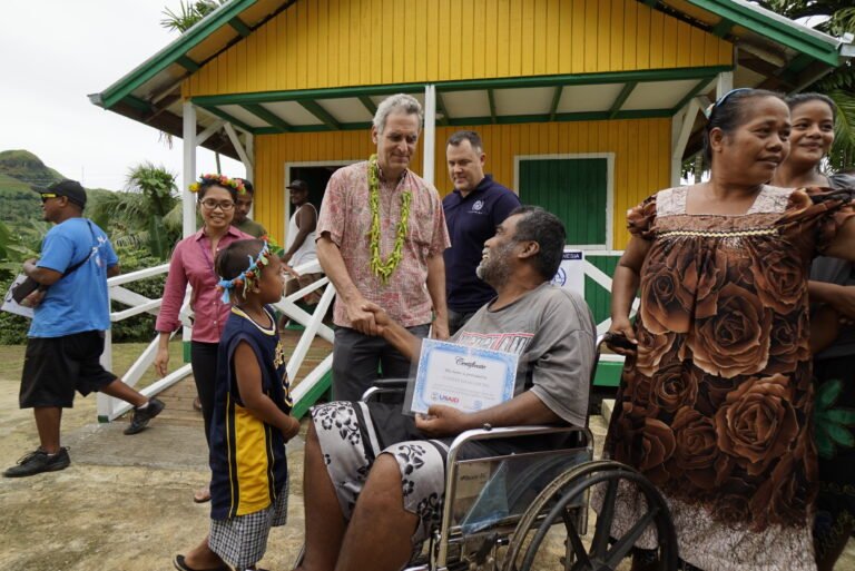 Man in wheelchair shakes hands with man, surrounded by community members and new resilient building in background.