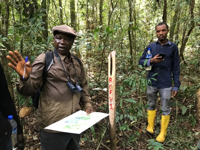 A man stands in the forest, listening intently to another man who is holding a paper and making a presentation.