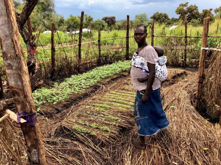 A woman with a baby on her back stands proudly in a fenced garden plot.