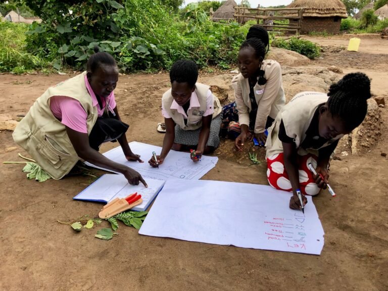 A group of women sits outside and draws on a large paper.