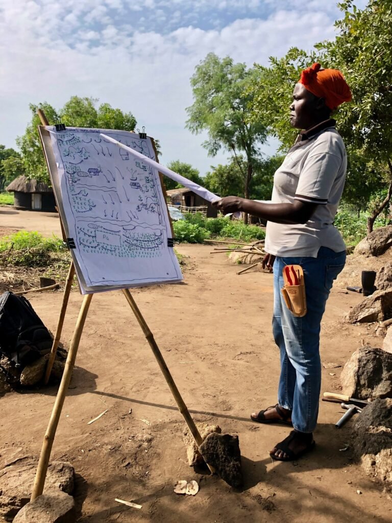 A woman stands outside and gestures towards an easel with a hand-drawn map on it.