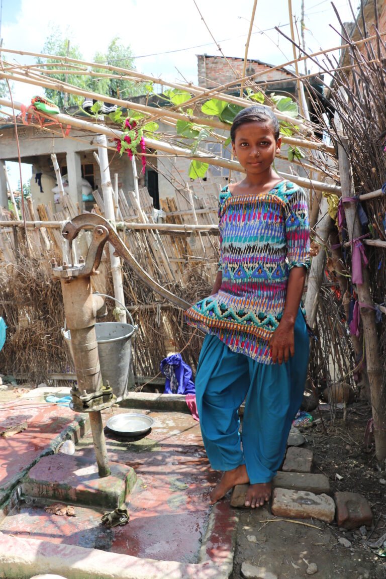 A young girl stands next to a water pump, looking at the camera.
