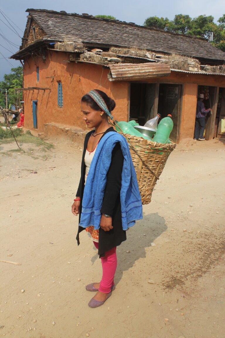 A young woman walks with heavy water jugs in a basket suspended from her back.