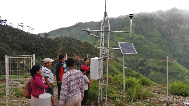 A group of people gather around the bottom of a weather station located on the slopes of a mountain.