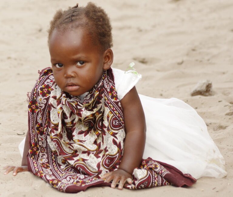 Small girl sitting on sandy ground and looking at the camera.