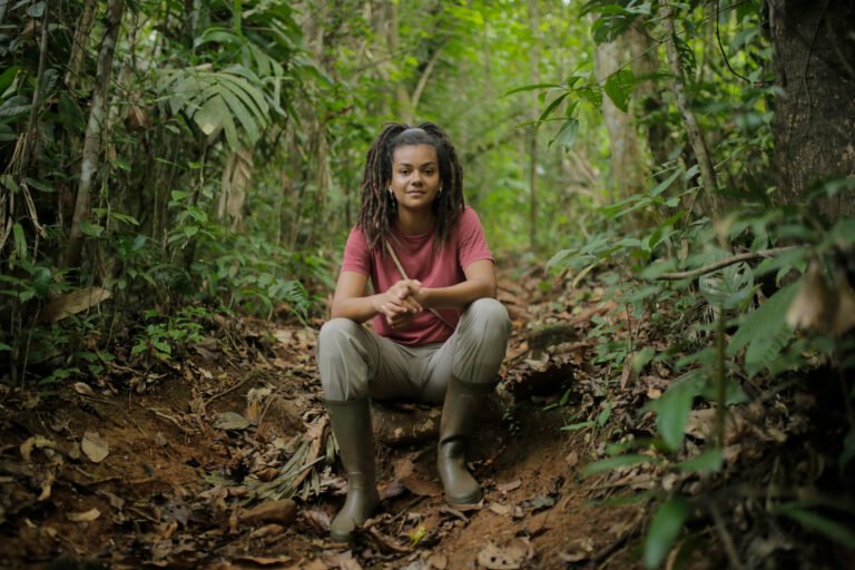 A woman sits in the middle of a jungle trail, looking at the camera.