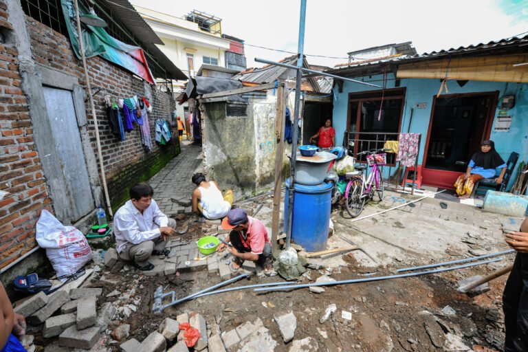 Workers install water management infrastructure behind houses in a small alleyway.