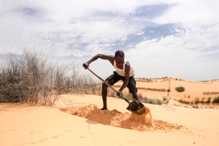 A man shovels sand in a desert area with sand dunes visible in the background.