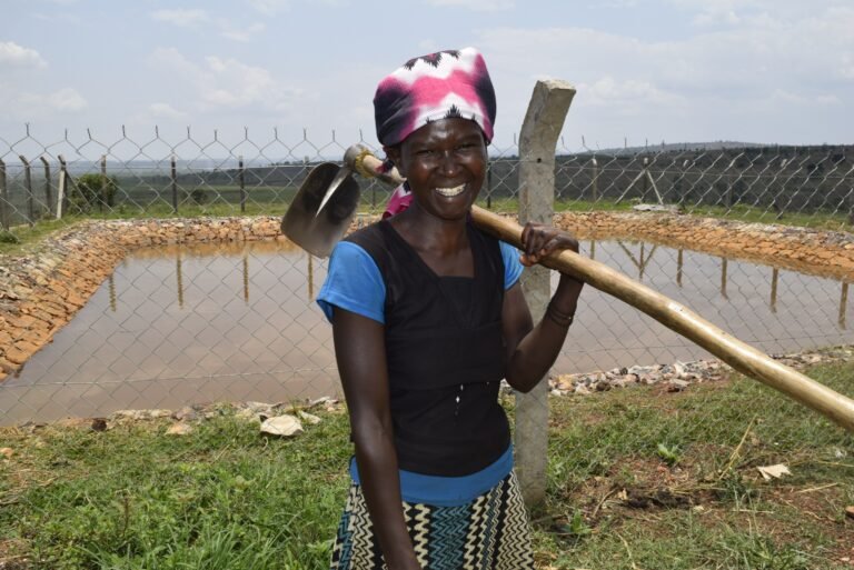 A woman holding a hoe stands in front of a fenced-in pond, smiling at the camera.