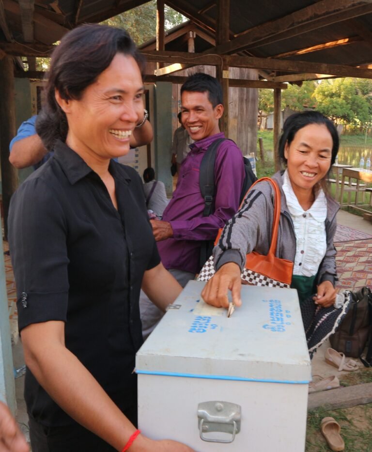 A woman places money into a metal lockbox held by another smiling woman.